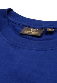 T-shirt in cotone blu con collo tondo, collare a costine e una piccola etichetta del marchio con testo dorato su sfondo nero.