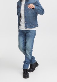 Džinsinis švarkas su šviesiai mėlyna spalva, sagų užsegimu ir priekinėmis kišenėmis, derinamas su tamsiai mėlynomis siaurų džinsų kelnėmis ir juodais bateliais su raišteliais.