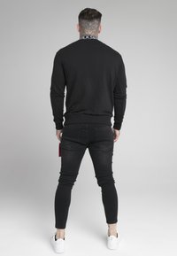 Man med nacktattoo som bär svart sweatshirt, svarta skinny jeans och vita sneakers, står vänd bort på grå bakgrund.