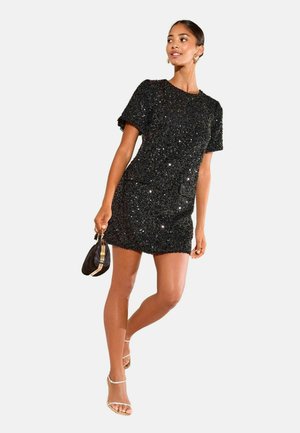 Schwarzes, mit Pailletten besetztes Kleid mit kurzen Ärmeln, taillierter Form und rundem Ausschnitt. Das Modell hält eine kleine schwarze Tasche und trägt Riemchen-High Heels.