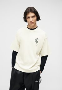 Νεαρός μεσαίου μήκους σκούρα μαλλιά, φοράει μαύρο μακρυμάνικο ρούχο πολλαπλών στρώσεων κάτω από ένα άσπρο μπλουζάκι με γραφικό τίγρη και μαύρο παντελόνι Adidas.