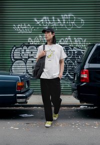 Tricou alb cu grafică verde, pantaloni largi negri, geantă gri și adidași verzi cu talpă albă, stând pe o stradă lângă mașini parcate.