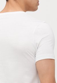 T-shirt blanc à manches courtes avec un col rond, texture lisse, présentant un logo en relief discret au dos près de l'encolure.