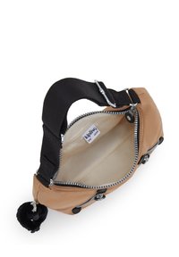 Beige kangas kott med svart rem, silverdragkedja och rund svart pom-pom charm. Öppen topp avslöjar en beige innervägg.