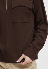 Brown pullover top with a zippered neck, featuring a front patch pocket, ribbed cuffs, and a relaxed fit. Soft, textured fabric.