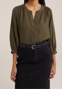 Olijfgroen blouse met knopen en een gestructureerde afwerking, met geplooide schouders en een ronde kraag, gecombineerd met een zwarte rok en riem.