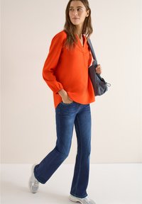 Femme marchant avec la main dans la poche, portant un chemisier orange, un jean bleu, des baskets blanches et un sac à bandoulière gris.