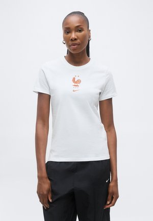 Žena v bielej tričku s ružovo-zlatým emblémom kohúta a čiernych nohaviciach Nike, stojaca pred jednofarebným bielym pozadím.