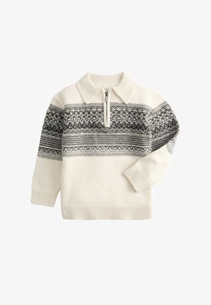 Jeune garçon regardant vers le bas sur sa gauche, portant un pull blanc à motifs, un pantalon en velours côtelé vert et des bottes noires sur un fond clair.