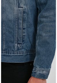 Denim jacket in light blue, featuring a classic cut, side pocket, and visible stitching. Cuffs are rolled with a clean finish.