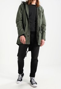 Parka verde con cappuccio sopra un maglione nero strutturato, abbinato a jeans neri e sneaker alte nere, con lacci e suole bianche.