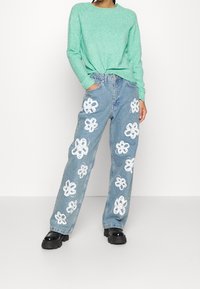 Hellgrüner Strickpullover, blaue Jeans mit weißen Blumenmustern, lockere Passform und schwarze Stiefel mit dicker Sohle.