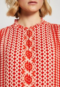 Blouse à motifs rouge et blanc avec un col rond, fermeture à boutons, et des designs géométriques et floraux élaborés sur un tissu léger.