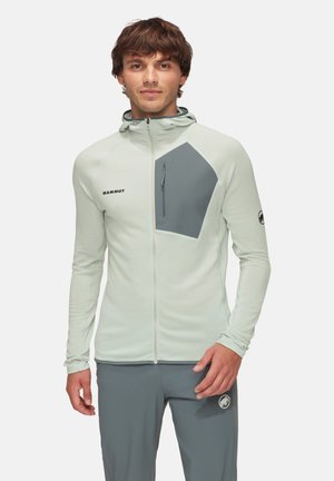 Jeune homme portant une veste zippée gris clair à capuche avec une poche poitrine gris foncé, assortie à un pantalon gris foncé.