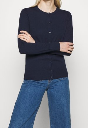 Person iført en knappet navyblå cardigan med lange ærmer og blå denimjeans, stående med armene over kors.