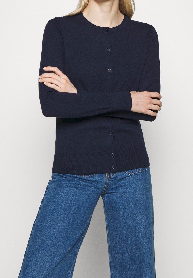 Personne portant un cardigan bleu marine boutonné à manches longues et un jean en denim bleu, debout les bras croisés.