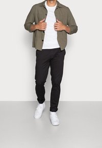 Camicia verde oliva con bottoni, t-shirt bianca, pantaloni neri a sigaretta e sneakers bianche. Outfit casual con una vestibilità rilassata e maniche arrotolate.