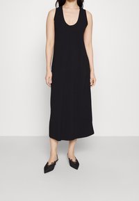 Schwarzes ärmelloses Kleid aus weichem Stoff, mit einem Rundhalsausschnitt und einem lässigen Schnitt, der bis zur Mitte der Waden reicht. Getragen mit spitzen schwarzen Schuhen.