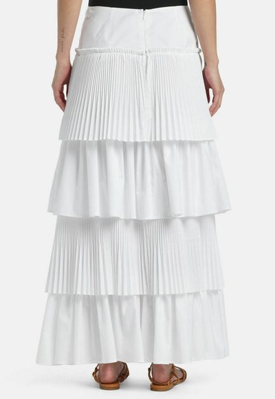 Set bagfra af person iført langt, hvidt, lagdelt nederdel med plisseringer og brune flade sandaler på en ensfarvet baggrund.