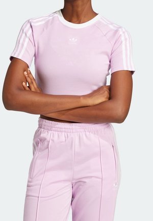 Person iført lyse lilla Adidas atletisk outfit med hvide striber på ærmer og bukser, arme krydset over brystet.