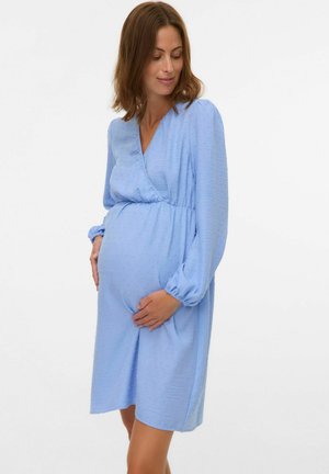 Zwangere vrouw in een lichtblauwe jurk met lange mouwen wiegt voorzichtig haar buik en kijkt met een zachte glimlach naar beneden.