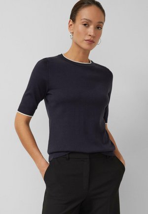 Femme portant un haut en maille à manches courtes bleu marine avec bordure blanche et pantalon noir, debout les mains dans les poches devant un fond uni.