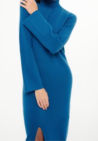 Långärmad klänning med hög hals i livlig teal, tillverkad av mjukt tyg, med en slits i sidan och avslappnad passform med vida ärmar.