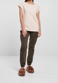 Kobieta ubrana w jasnoróżową bluzkę z krótkim rękawem, ciemno-brązowe spodnie jogger z kieszeniami i futrzane brązowe klapki na otwartym palcu, stojąca na gładkiej białej podłodze.