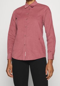 Langarm, overhemd met kraag in gedempt rood corduroy. Heeft een knoopsluiting aan de voorkant, een lichte kromming aan de onderkant en subtiele logo borduring nabij de onderrand.