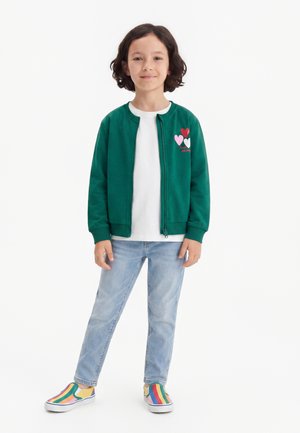 Barn med kort mørkt hår har en grøn jakke med lynlås og hjerte-design, hvid skjorte, lyseblå jeans og farverige stribede slip-on sko.