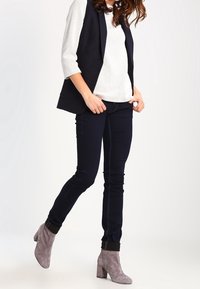 Pantalones slim-fit azul marino combinados con una blusa blanca texturizada y un chaleco oscuro, complementados con botas de ante gris de tobillo con detalles en el dobladillo.