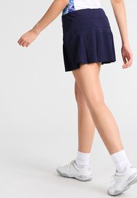 Falda deportiva plisada de color navy hecha de una tela elástica, con una cinturilla elástica. El modelo lleva zapatillas deportivas y calcetines blancos.