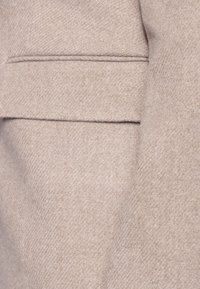 Beige ylletyg med fiskbensmönster, slät textur och en fickdetalj som visar subtila sömmar och mjuk fall.