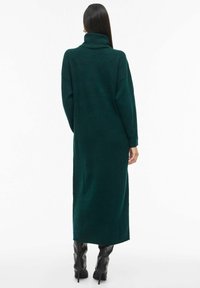 Garšs zaļš turtleneck kleita, izgatavota no mīksta adījuma. Tam ir brīvs piegriezums un pilna garuma piedurknes, apvienota ar melniem augstiem zābakiem.