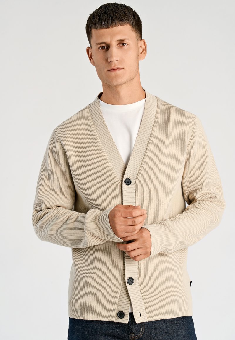 Beige geribbelde cardigan met een V-hals, een enkele zwarte knoopsluiting en lange mouwen, gedragen over een wit overhemd en gecombineerd met een donkere spijkerbroek.