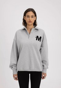 Szary sweter z kapturem i kołnierzykiem, z haftowanym czarnym logo "M" na lewej piersi oraz długimi, luźnymi rękawami.