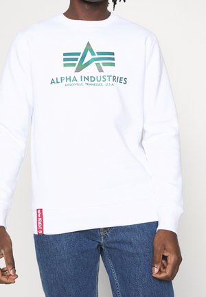 Czarna osoba ubrana w biały sweter Alpha Industries z zielonym logo i napisem, w połączeniu z niebieskimi dżinsami, stojąca na jasnym tle.