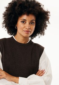 Maglione senza maniche marrone sopra una maglietta a maniche lunghe bianca con coste. Il modello ha capelli neri ricci e indossa orecchini a cerchio in argento.