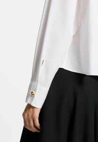 White long-sleeve shirt with a cuff featuring a gold button detail; smooth fabric with a slight sheen; black skirt with pleats.