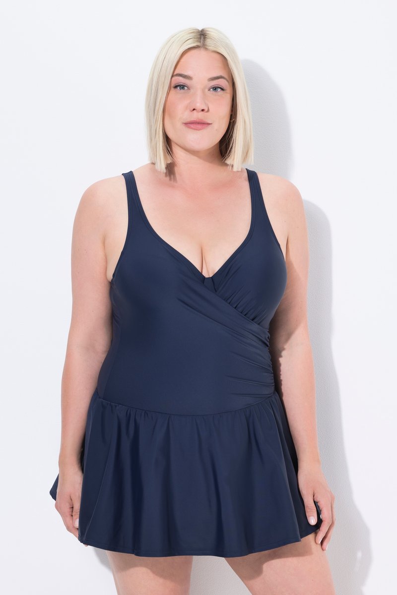 Donna bionda che indossa un costume da bagno senza maniche blu navy con scollo a V e dettaglio arricciato sul lato, in piedi su sfondo bianco.