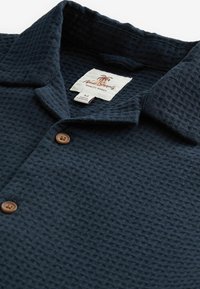 Polo de color azul marino hecho de tela texturizada con cuello. Presenta dos botones marrones y una etiqueta con el logo de una palmera y talla M.
