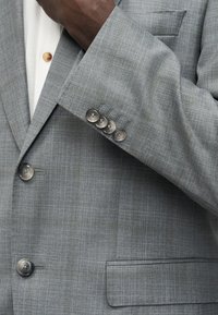 Gros plan sur une personne portant une veste de costume à carreaux gris avec quatre boutons aux poignets et une chemise blanche avec un bouton en bois visible.