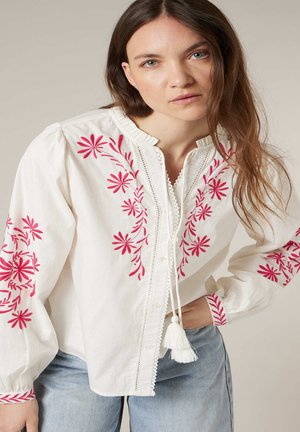 Jeune femme portant un chemisier blanc avec une broderie florale rose et des liens à pompons, associée à un jean bleu clair, posant avec une main sur la hanche.