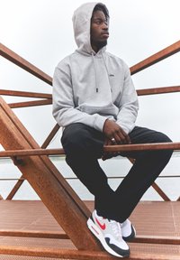 Grå hoodtröja med en framficka och dragsko, svarta byxor och vita sneakers med röda och svarta accenter, sittande på en metallstruktur.