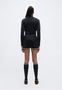 Άτομο που στέκεται με την πλάτη στην κάμερα, φορώντας μαύρο αθλητικό μακρυμάνικο τοπ, μαύρο σορτς, κάλτσες μέχρι το γόνατο και παπούτσια ποδοσφαίρου σε απλό φόντο.