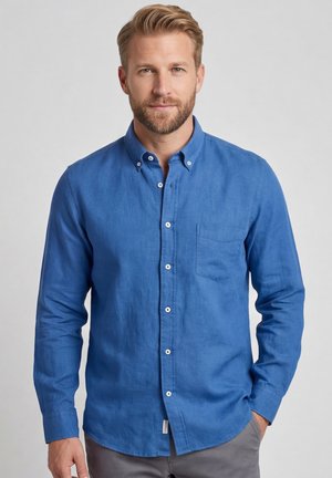 Man met baard die een blauw overhemd met knopen en lange mouwen en grijze broek draagt, staand tegen een effen achtergrond.