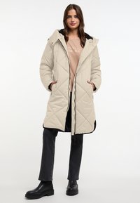 Beige gewatteerde lange jas met een capuchon, ritsluiting en zwarte accenten. Draagt over een licht shirt en gecombineerd met zwarte broek en robuuste laarzen.
