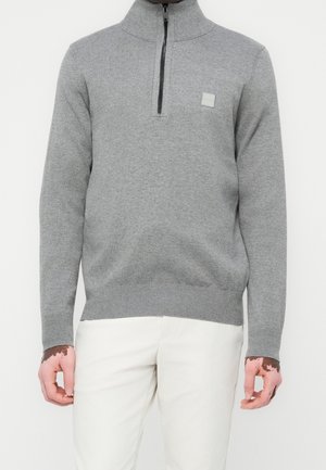 Mann trägt einen hellgrauen Pullover mit Viertel-Reißverschluss und weiße Hose, Hände sichtbar mit Vitiligo auf der Haut.
