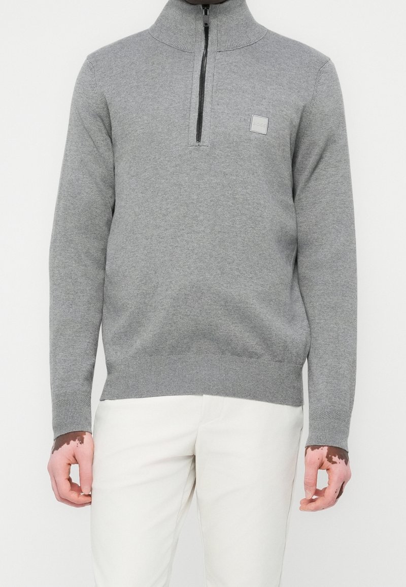 Homme portant un pull gris clair à fermeture éclair au quart et un pantalon blanc, les mains visibles montrant du vitiligo sur la peau.