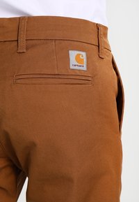 Bruine katoenen broeken met een achterzak, voorzien van een Carhartt-logo patch. De stof heeft een gladde textuur met een subtiele diagonale weving.